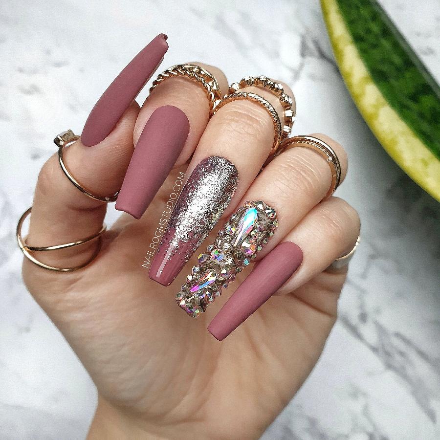 Stylish matte mauve nails with a gold chrome ombre effect and a crystal accent on one nail, displayed on a white hand with jewelry