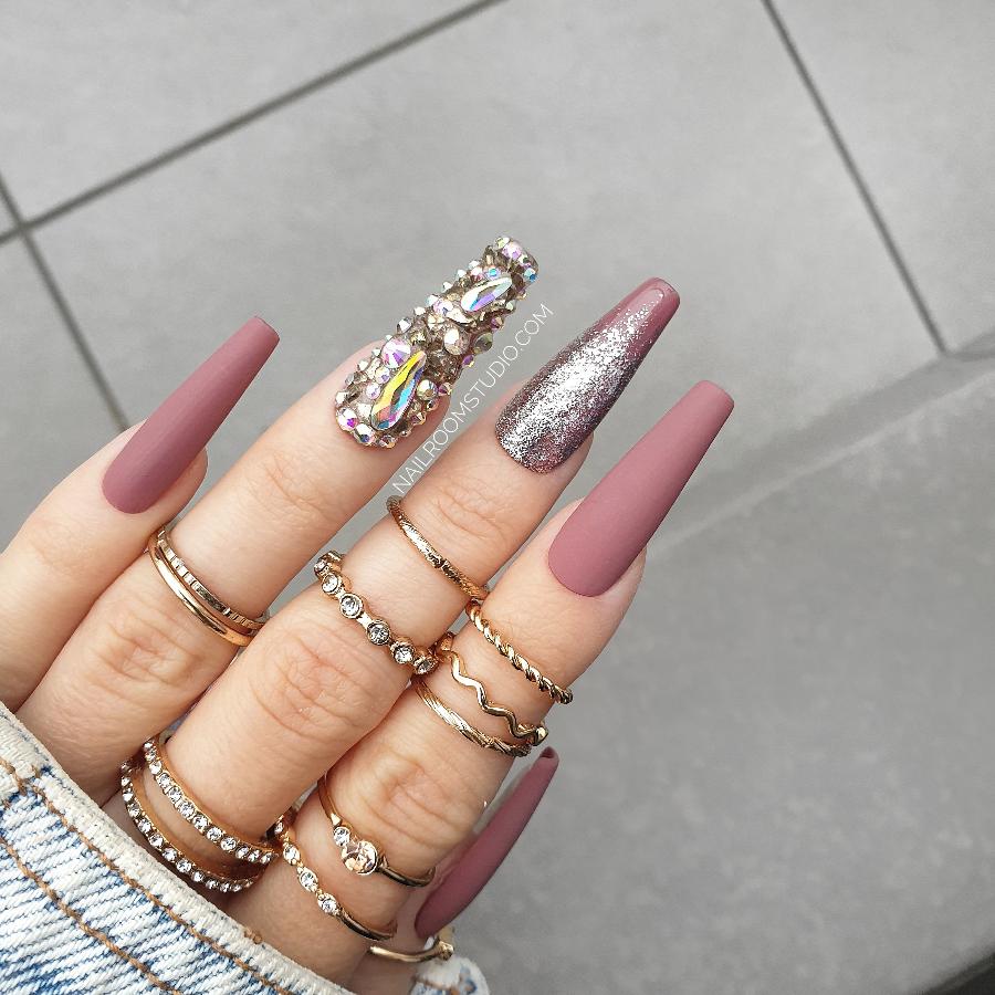 Matte mauve coffin nails with a gold chrome ombre effect, featuring a crystal-accented nail, displayed on a white hand with rings