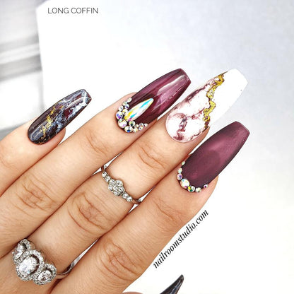 Long coffin nails in burgundy with black and white marble accents, featuring gold crystals by Nail Room Studio