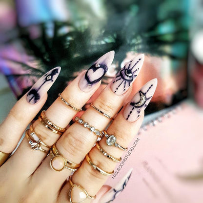 Long stiletto nude nails with hand-drawn symbols, including a heart, moon, and eye, for a whimsical look by Nail Room Studio