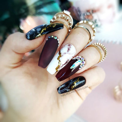 Burgundy coffin nails in a matte finish, accented with marble patterns and gold crystals by Nail Room Studio