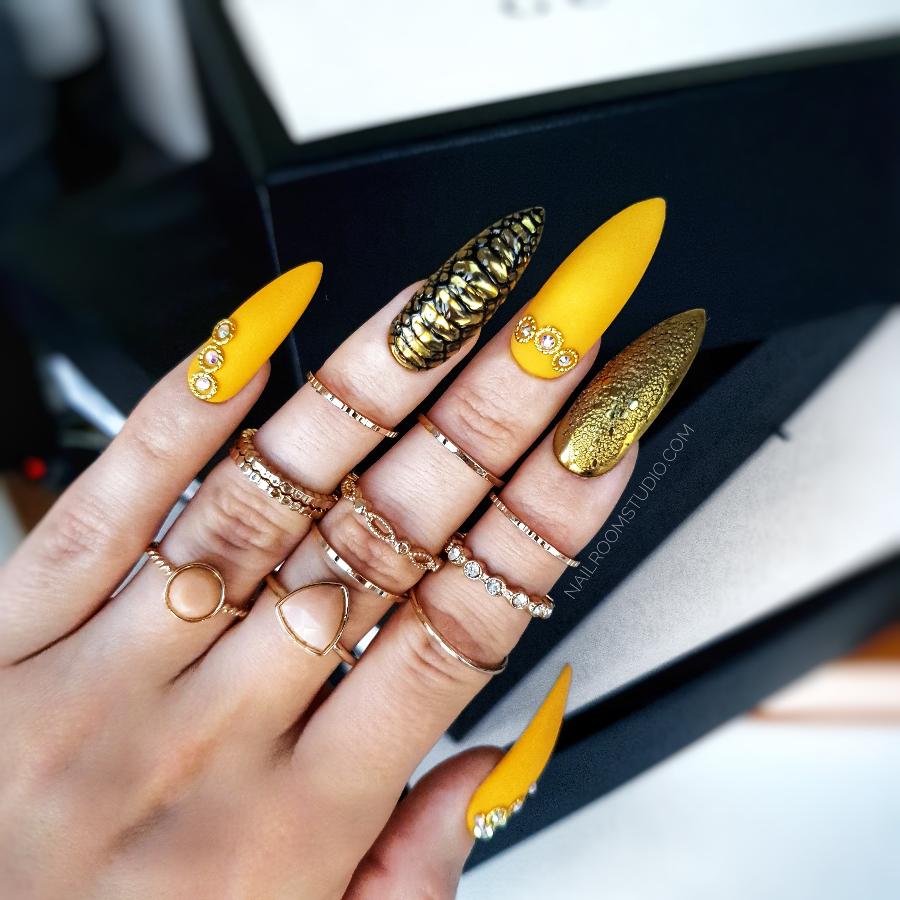 Yellow-orange matte nails inspired by dragon and snake skin, adorned with oriental metal accents, featuring a French Bollywood design