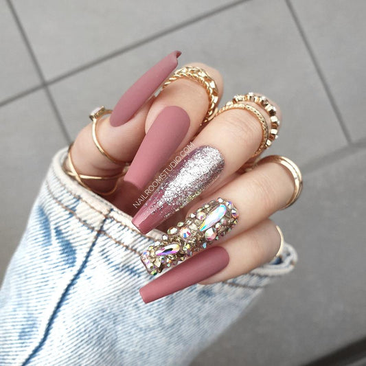 Elegant mauve coffin nails in a matte finish with gold chrome ombre tips and one crystal-accented nail, shown on a hand with rings