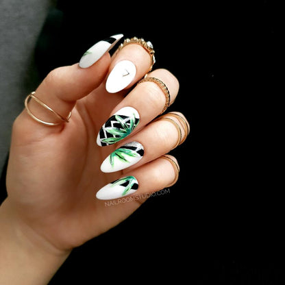 White nails with black and green jungle-themed botanical designs and geometric details, displayed on hands with rings for a contemporary style