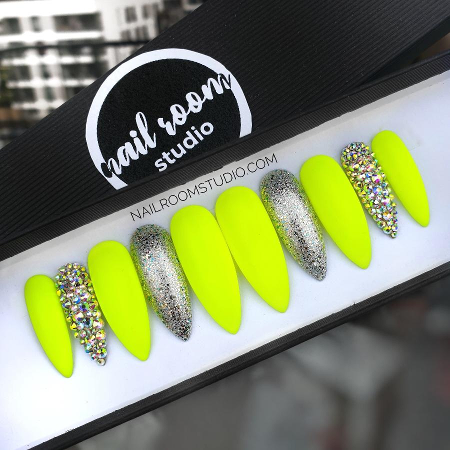 Vibrant neon yellow press-on nails in long stiletto shape, matte finish with silver chrome accents on one nail and full opalescent silver Swarovski crystals on another, perfect for summer, displayed on hands