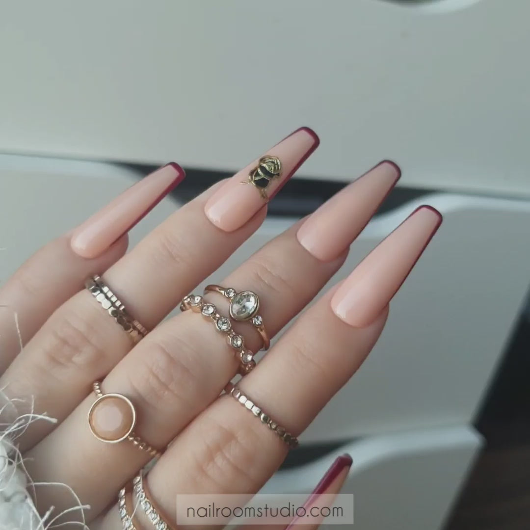 Peachy nude glossy nails with burgundy french tips, black roses, and gold accents, creating a chic witchy and mortal look by Nail Room Studio