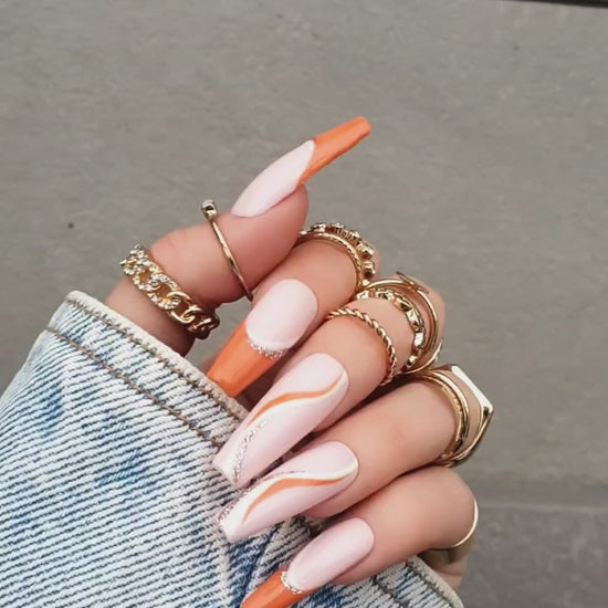 Elegant powder pink Press-On nails with orange French tips, decorated with delicate gold and orange wave accents, showcased on a hand with long nails