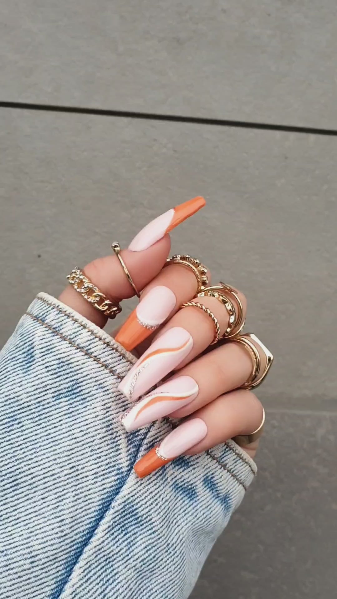 Elegant powder pink Press-On nails with orange French tips, decorated with delicate gold and orange wave accents, showcased on a hand with long nails