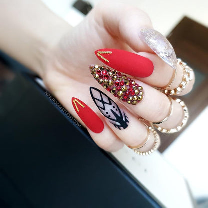 Deep red femme fatale nails with lace accents adorned with gold and red Swarovski crystals featuring a bold chrome gold finish for a seductive daring look

